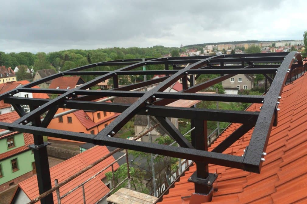 Czarna stalowa konstrukcja zadaszenia z łukowymi krokwiami na dachu budynku, w tle panorama miasta